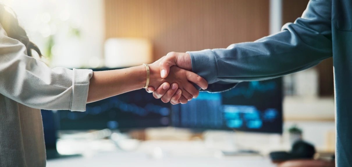 Two people shaking hands in front of a computer, with a blurred background